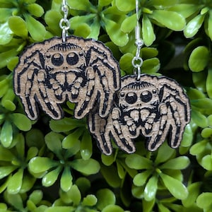 May include: A pair of wooden earrings featuring a black and white illustration of a jumping spider. The earrings are hanging from silver hooks.
