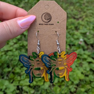 May include: Colorful wooden bee earrings with blue, green, yellow, and red accents. The earrings are hanging from silver hooks and attached to a brown tag with the text "WICKED FOREST DESIGNS".
