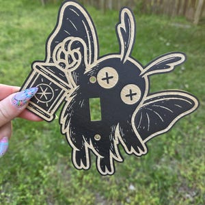 May include: Black and tan light switch cover featuring a cartoon moth holding a lantern. The moth has cross-shaped eyes and a furry body. The lantern has a star design. The switch cover is made of wood.