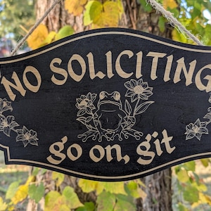 May include: Black wooden sign with the words "NO SOLICITING" and "go on, git" in a light brown colour. A frog and floral design is in the centre. The sign is hanging from a tree.