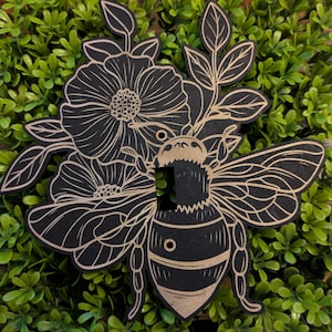 May include: Black and tan wooden light switch cover featuring a bee design with floral accents. The bee is centered with detailed wings and body stripes. The flowers and leaves are intricately carved, creating a nature-inspired decorative element.