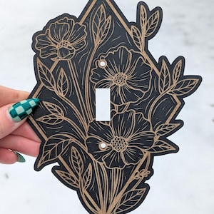 May include: Black and gold floral light switch cover. The cover has a diamond shape with a floral design. The flowers and leaves are gold, and the background is black. The light switch opening is white.