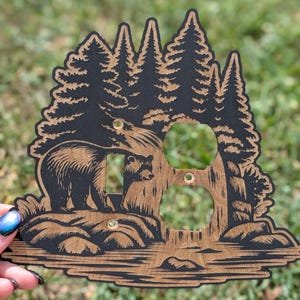 Könnte beinhalten: Holz-Lichtschalterabdeckung mit einem Bären und einem Jungtier in einer Waldszene. Das Design umfasst Bäume, Felsen und Wasser, mit zwei Steckdosenlöchern. Das Kunstwerk ist in Schwarz- und Brauntönen gehalten und erzeugt eine rustikale Ästhetik.