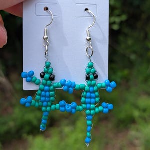 May include: A pair of beaded earrings featuring two lizard-like figures. The earrings are made with teal and blue beads, with black bead eyes and silver-colored ear wires. The figures are connected at the hands.