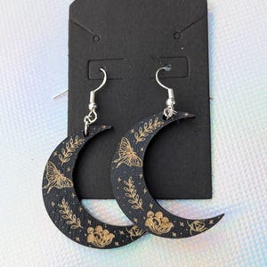 May include: A pair of crescent moon earrings. The black earrings feature a gold butterfly, floral, and leaf design. The earrings hang from silver-colored hooks and are displayed on a black card.