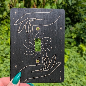 May include: Black light switch cover with a celestial design. The cover features two hands reaching towards a swirling pattern, with small stars and dots. The design is in a gold color, and the cover has two green circles.