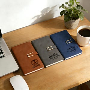 Puede incluir: Tres cuadernos personalizados en marrón, gris y azul, cada uno con un cierre dorado. El cuaderno marrón tiene el nombre "Alexander" y un logotipo. El cuaderno gris tiene el nombre "Heather" y la fecha. El cuaderno azul tiene el nombre "Michael".