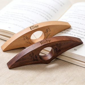 Puede incluir: Dos sujetapáginas de madera, uno claro y otro oscuro, con grabados florales. El sujetapáginas claro tiene grabado el nombre "Elise", mientras que el oscuro tiene "Ruby". Ambos tienen una abertura circular para el dedo.