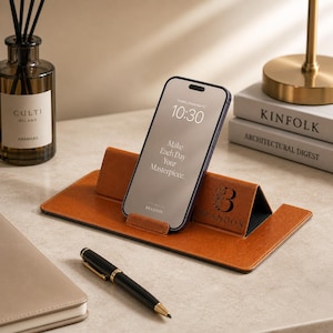 Può includere: Un supporto per telefono in pelle marrone con uno smartphone che mostra l'ora 10:30 e il testo "Make Each Day Your Masterpiece". Il supporto è personalizzato con la lettera "B" e il nome "BRANDON". Sono visibili anche una penna nera e un taccuino.