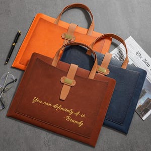 Peut inclure: Trois porte-documents en cuir orange, marron et bleu marine. Chacun a une poignée, une sangle en cuir avec un fermoir doré et une poche. Le porte-documents marron porte l'inscription "You can definitely do it. -Brandy."