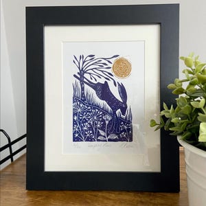 May include: Framed art print featuring a blue hare leaping near a tree and flowers, with a golden sun in the background. The print is set against a white mat and framed in a black frame. The artwork is signed.