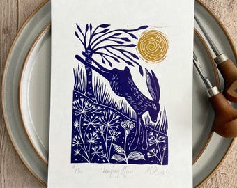 May include: A rectangular art print on white paper featuring a dark blue linocut illustration of a hare leaping through a field of flowers under a tree and a golden yellow sun. The print is titled Leaping Hare and numbered 9/30.