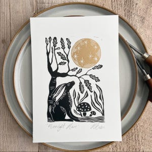 May include: A white rectangular print featuring a black ink illustration of a hare sitting under a tree with a gold moon and stars above. Two mushrooms are visible near the hare. The text 'Moonlight Hare' and a signature are at the bottom.