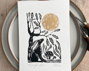 May include: A white rectangular print featuring a black ink illustration of a hare sitting under a tree with a gold moon and stars above. Two mushrooms are visible near the hare. The text 'Moonlight Hare' and a signature are at the bottom.