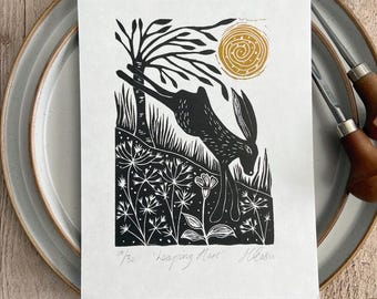 May include: A black and white linocut print on paper featuring a leaping hare, a tree, and a golden sun. The print includes the handwritten text 10/30, Leaping Hare, and a signature at the bottom. The artwork is centered on a plate.