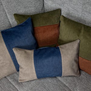 Könnte beinhalten: Eine Ansammlung von vier rechteckigen Dekokissen mit Color-Block-Designs in den Farben Olivgrün, Dunkelblau und Taupe. Die Kissen sind in einer Gruppe angeordnet und zeigen glatte Stofftexturen sowie saubere, gerade Nähte zwischen den Paneelen.