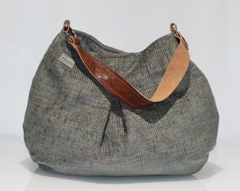 May include: A large, textured gray and brown handbag with a brown leather strap. The bag has a rounded shape and a single shoulder strap. The strap is attached to the bag with metal rings. A small tag is sewn on the side of the bag.