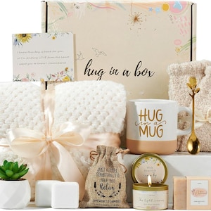 May include: A gift box labeled "hug in a box" filled with self-care items. Includes a white blanket, mug with "Hug in a Mug", candle, soap, and a succulent. A card reads "I know this day is hard for you..."