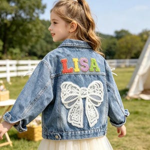 May include: Light blue denim jacket with a large white lace bow on the back. The word "LISA" is embroidered across the upper back in colorful letters. The jacket has a classic collar and button closures.