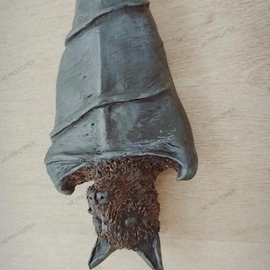 Pode incluir: Uma escultura de morcego cinza escuro com um corpo texturizado e rosto detalhado, pendurada de cabeça para baixo. As asas do morcego estão dobradas e seu rosto tem olhos e orelhas visíveis. A escultura é feita de um material escuro.