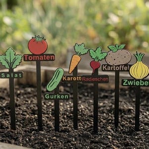 Könnte beinhalten: Schwarze Metall-Gartenstecker mit farbenfrohen Gemüseillustrationen. Die Stecker zeigen eine Tomate, Karotte, Gurke, Radieschen, Kartoffel, Zwiebel und Salat. Jeder Stecker trägt den Namen des Gemüses.