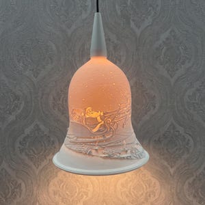 Könnte beinhalten: Eine weiße, glockenförmige Porzellan-Hängeleuchte mit einem durchscheinenden Lithophanie-Design, das den Weihnachtsmann in einem von Rentieren gezogenen Schlitten zeigt und von innen beleuchtet wird, um die winterliche Szene warm zu erhellen.