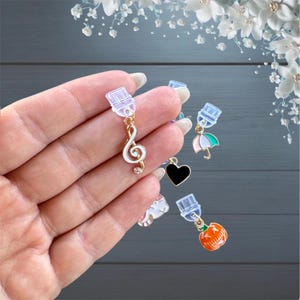 Peut inclure: Une collection de breloques décoratives, dont une clé de sol blanche, un cœur noir, un parapluie rose et vert et une citrouille orange. Chaque breloque est attachée à une pince transparente avec une boucle en métal doré.