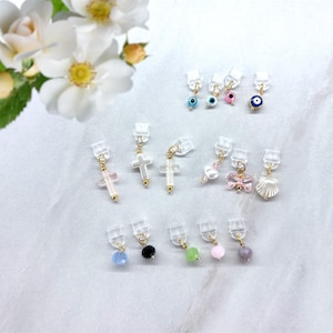 Op de afbeelding: Een verzameling clip-on oorbellen met verschillende ontwerpen. Stijlen zijn onder meer kruisen, strikken, schelpen en kralen in kleuren als roze, wit, blauw en zwart. De oorbellen hebben goudkleurige accenten en doorzichtige clips.