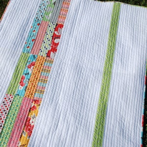 May include: A handmade quilt with a white, vertically-striped background and colorful patchwork strips. The patchwork includes floral and patterned fabrics in various colors. A green patterned border and a green stripe accent the quilt.