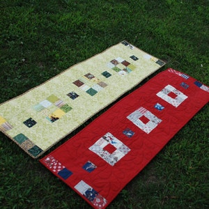May include: Two handmade table runners on green grass. One is light green with a floral pattern and colorful square patches. The other is red with square patches and quilted details. Both have decorative borders.