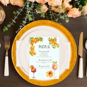 Op de afbeelding: Tafelsetting met een goudgerande bord, bestek en een menukaart. De menukaart toont sinaasappel illustraties en het woord "MENU". Bloemschikkingen en een brandende kaars dragen bij aan de sfeer. De setting staat op een donkere houten ondergrond.