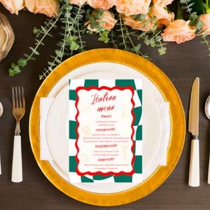 Op de afbeelding: Een Italiaanse menukaart met een rode en witte rand ligt op een wit bord, bovenop een goudgerande oplader. Bestek met witte handvatten en een gouden vork zijn rond het bord gerangschikt. Perzikroze bloemen en groen decoreren de donkere houten tafel.