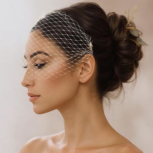 May include: A bridal veil with a net design, covering the face. The veil is secured with a pearl and a floral hair accessory. The hair is styled in a bun. The background is a soft, neutral color.