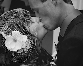 May include: A black and white photo of a couple kissing at their wedding. The bride is wearing a white veil with a flower on it. The groom is wearing a black suit and tie.