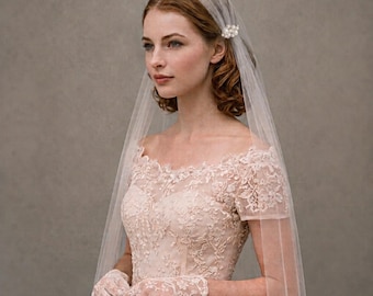 May include: A pale pink off-the-shoulder dress featuring intricate floral lace embroidery, short sleeves, and a sweetheart neckline. The ensemble includes matching lace gloves and a sheer tulle veil secured with a small white pearl hair accessory.