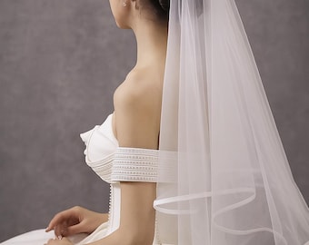 Könnte beinhalten: Ein weißer Brautschleier mit einem mehrschichtigen Design und einem Satintrimm. Der Schleier wird mit einem weißen schulterfreien Brautkleid kombiniert. Das Haar der Braut ist zu einem Dutt frisiert.
