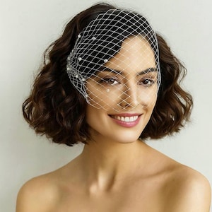 May include: A woman with short, dark brown hair smiles, wearing a white, net-like veil adorned with small, round pearls. The veil covers the top half of her face, creating a delicate and elegant look. The background is a plain, light color.