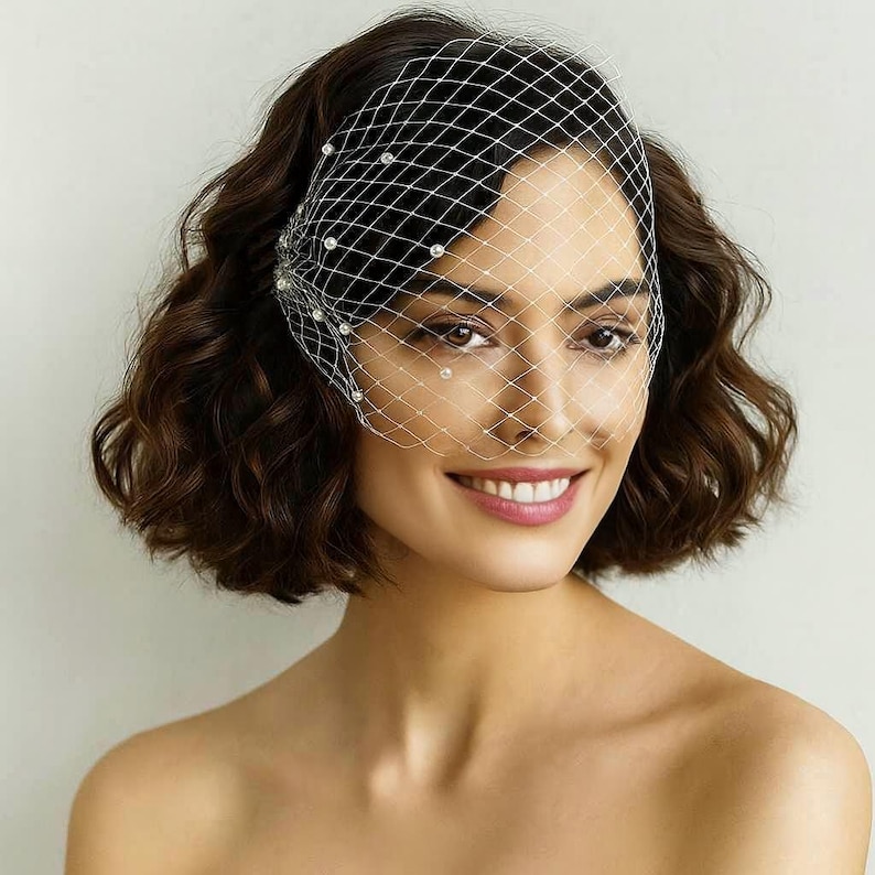 May include: A woman with short, dark brown hair smiles, wearing a white, net-like veil adorned with small, round pearls. The veil covers the top half of her face, creating a delicate and elegant look. The background is a plain, light color.