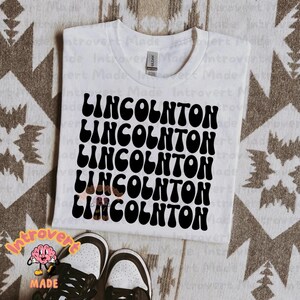 May include: White t-shirt with the name "LINCOLNTON" repeated five times in a black, retro, wavy font.
