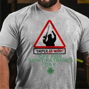 Może przedstawiać: Szary T-shirt z czerwono-białym znakiem ostrzegawczym, na którym widnieje napis "Sniper at Work Off for Saint Patrick's Day". Znak przedstawia sylwetkę osoby trzymającej karabin. Pod znakiem znajduje się zielona koniczyna z tarczą w środku.