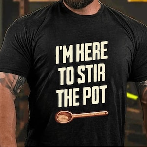 以下が含まれることがあります： 白い文字で「I'm here to stir the pot」と書かれた黒いTシャツ。テキストの下に木製のさじが描かれています。
