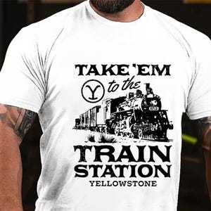 Op de afbeelding: Wit T-shirt met een zwart-witte afbeelding van een trein en de tekst "TAKE 'EM to the Y TRAIN STATION YELLOWSTONE".