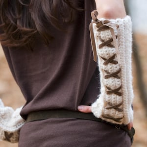 Könnte beinhalten: Ein Paar cremeweiße, gehäkelte Armstulpen mit strukturiertem Strickmuster und brauner Band-Schnürung, die sich über die gesamte Länge kreuzt und an den oberen Rändern zu kleinen Schleifen gebunden ist, um den Sitz an den Unterarmen zu sichern.
