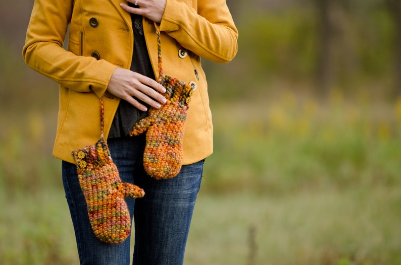 May include: A pair of crocheted mittens in shades of orange, brown, and yellow, with a button detail. The mittens are hanging from a yellow jacket.