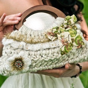 May include: A white crocheted handbag with a wooden handle and a floral fabric bow. The bag has a round mirror inside and a crocheted flower embellishment.