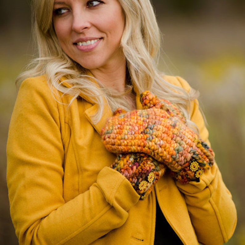 May include: A woman wearing a yellow jacket with a pair of colorful crocheted fingerless gloves. The gloves have a textured pattern and are decorated with small yellow flower embellishments.