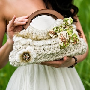 May include: Cream-colored, hand-knitted handbag with a wooden handle. The bag features a large, decorative flower and a patterned ribbon with a fairy design. The bag is embellished with gold trim and small beads.