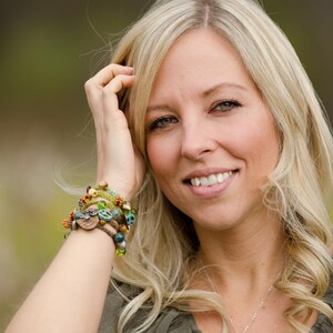 May include: A multi-strand beaded bracelet featuring a mix of green, brown, and turquoise beads, including small wooden accents, a peace sign charm, and a small owl-shaped charm, worn on a wrist with a green top and a silver chain necklace.