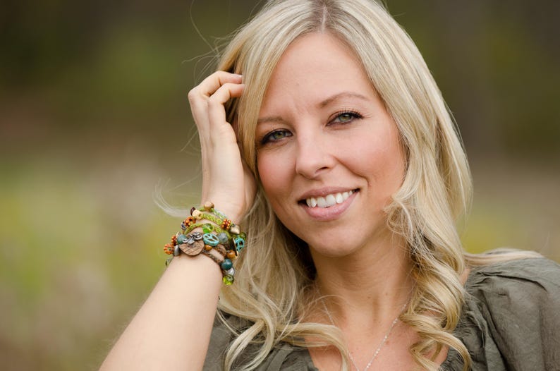 May include: A multi-strand beaded bracelet featuring a mix of green, brown, and turquoise beads, including small wooden accents, a peace sign charm, and a small owl-shaped charm, worn on a wrist with a green top and a silver chain necklace.