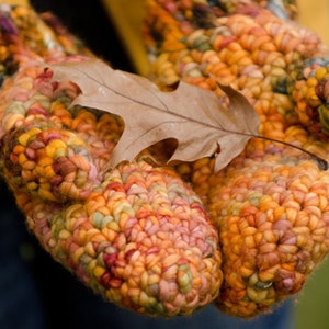 May include: A pair of colorful crocheted mittens with a brown leaf resting on top. The mittens are made with a variety of shades of orange, yellow, pink, and red yarn.